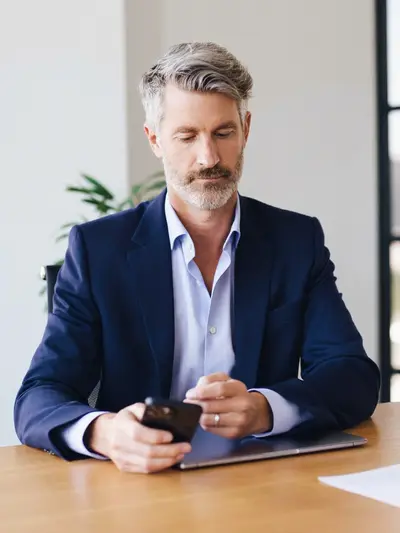 Sócio de escritório lendo uma mensagem no celular à mesa de trabalho
